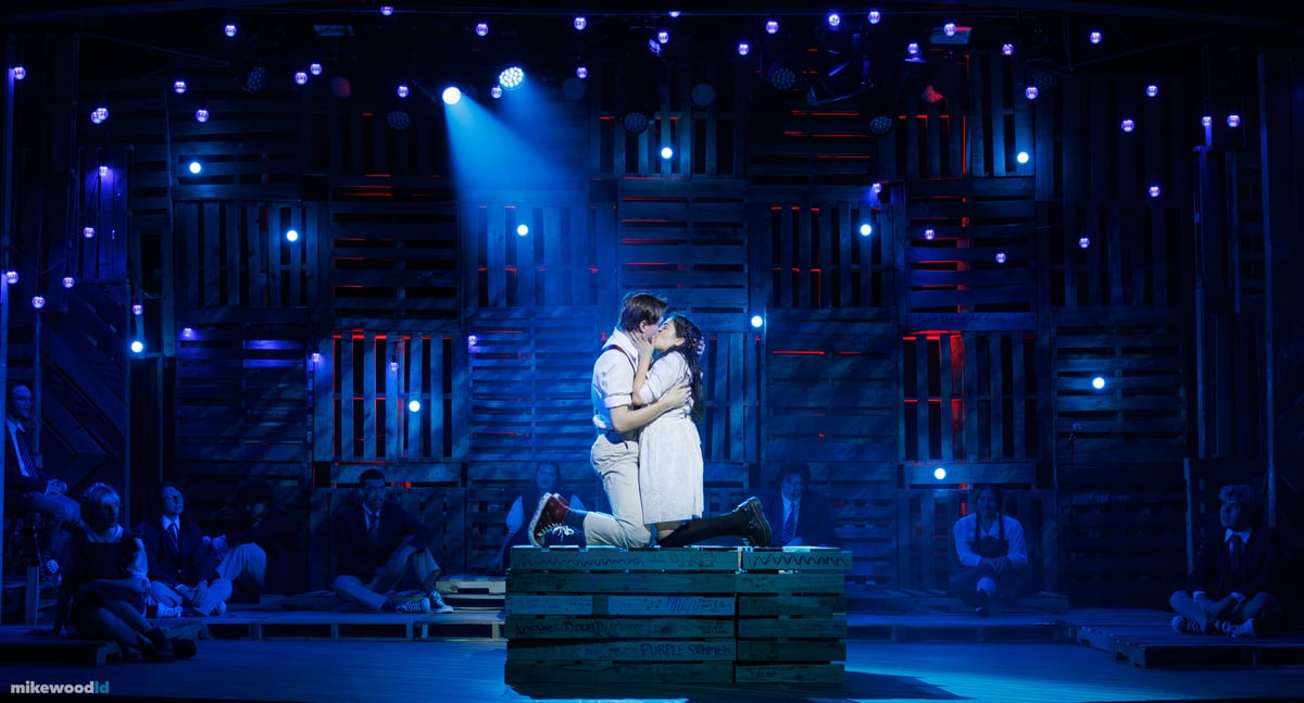 A young couple kneels and embraces atop a pallet crate in a tight blue spotlight, the ensemble seated in the dark around them and the pallet wall dotted with blue and red practicals.
