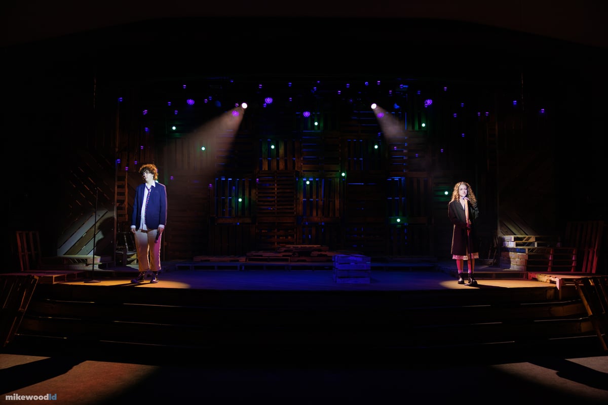 A young man and a young woman stand isolated far apart, each in a tight top special, the dark pallet wall behind them speckled with blue, green and violet pinlights.