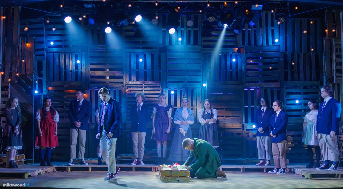 The company lines the stage watching a man in a green suit kneel over a flower-strewn coffin at center, the pallet wall behind washed in cool blue.