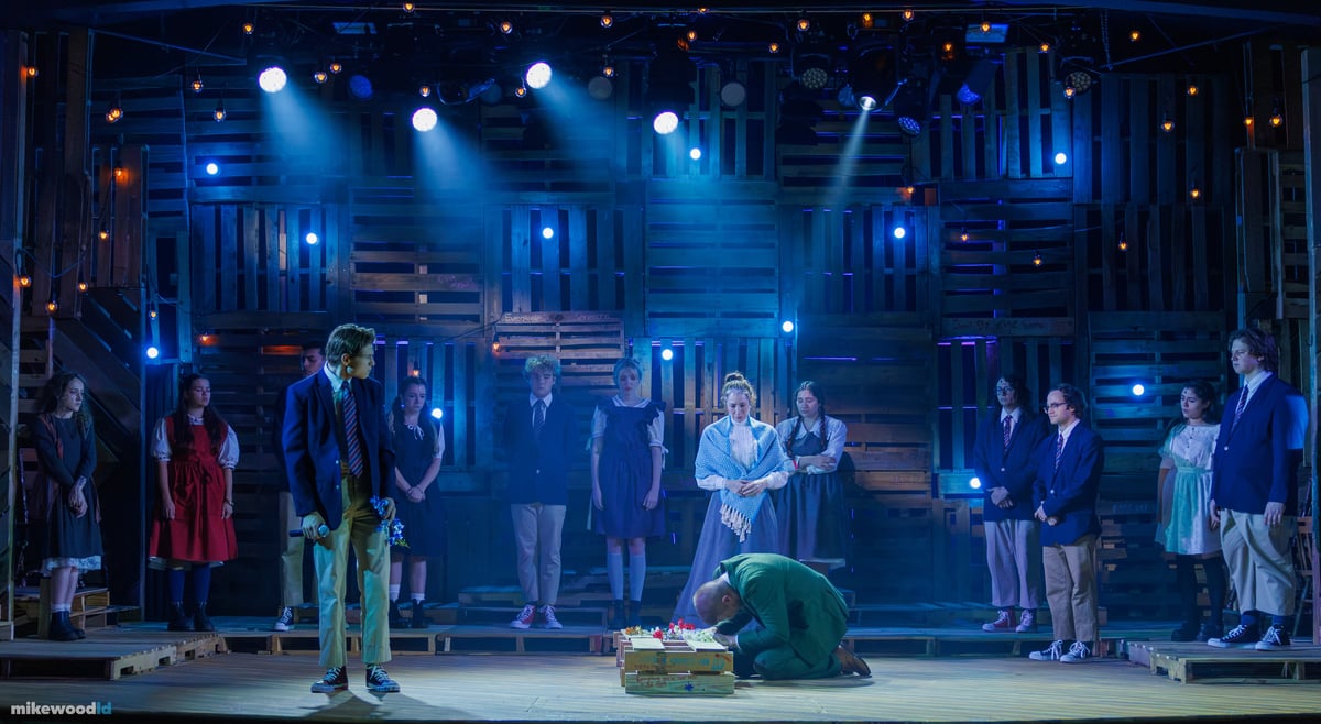A boy holding flowers faces a man kneeling at a small coffin as the company watches from the shadows in a cool blue wash.
