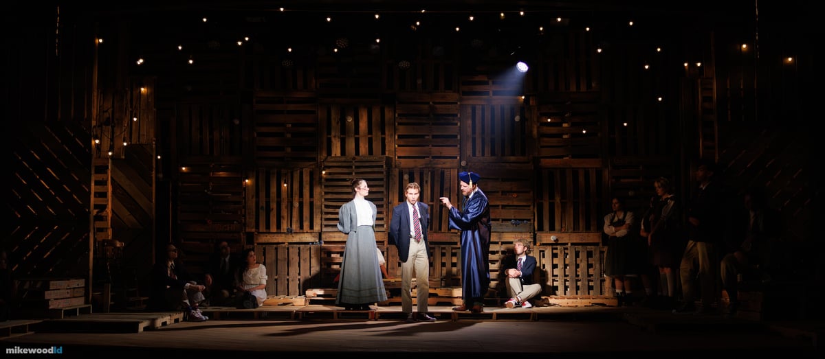 In a warm pool of light, a woman and a young man face a figure in cap and gown at center as the ensemble looks on from the dark pallet platforms.