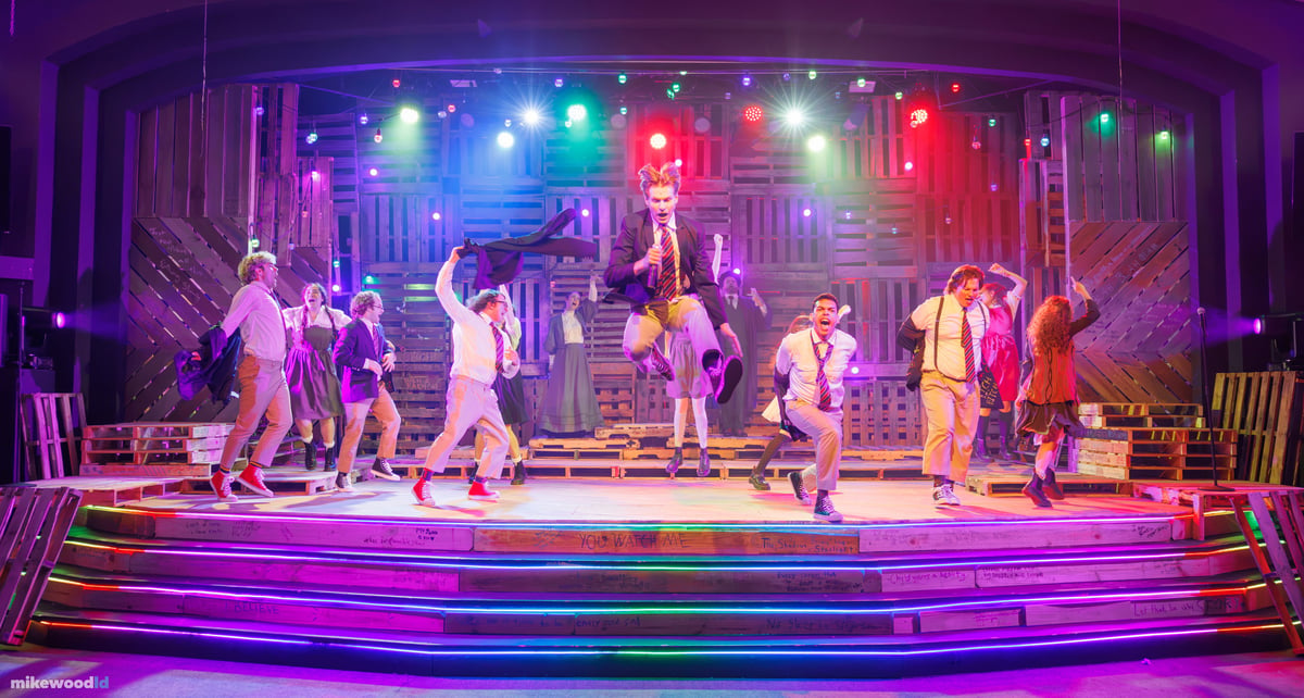 The full company breaks into dance, a young man airborne at center with a microphone, under a vivid multicolor wash with rainbow LED striping along the steps.