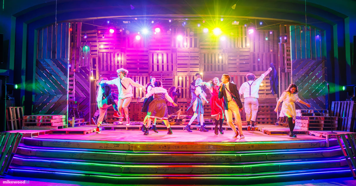 The company scatters into an exuberant dance across the stage under a saturated magenta and yellow wash, rainbow LED striping running along the step edges.