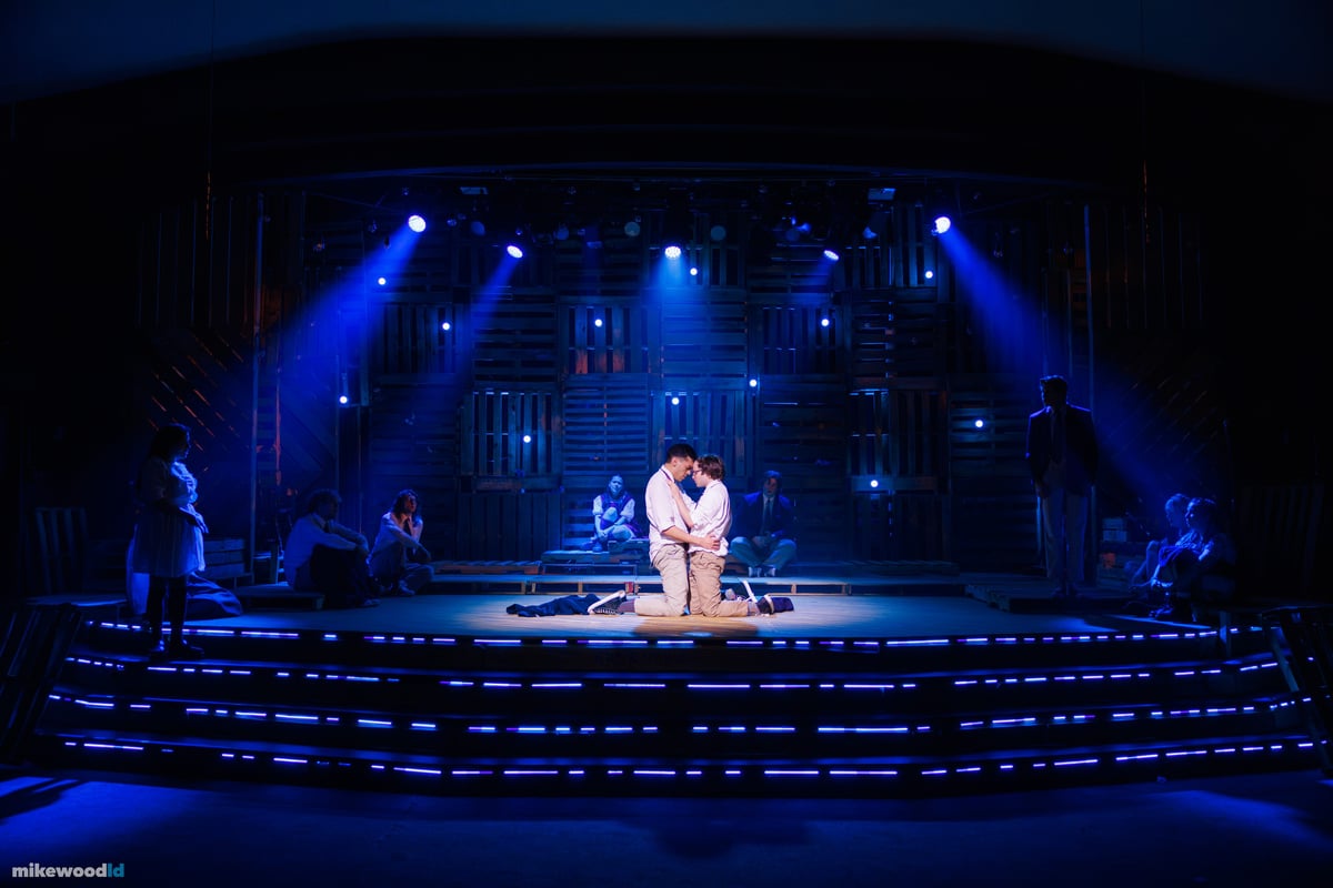 A young couple kneels together in a close embrace in a tight blue special, the ensemble watching from the shadows as blue LED striping dashes across the steps.