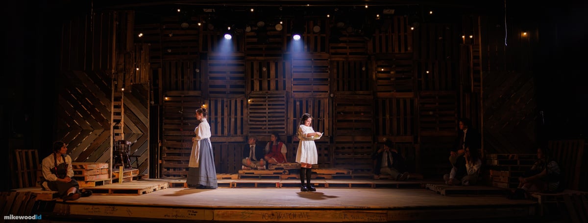 In a tight warm special, a young woman in white reads aloud at center as another in a grey dress faces her, the ensemble scattered across the dim pallet stage.