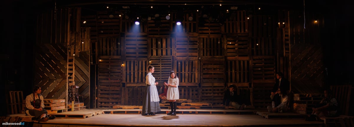 Two young women stand talking at center under a soft top light, the pallet wall washed warm amber and the ensemble seated in the surrounding dark.