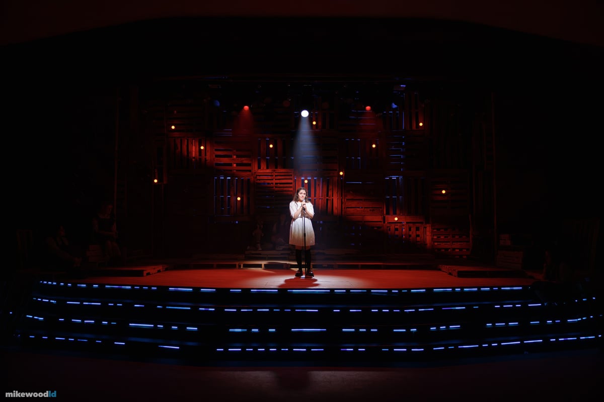 A young woman stands alone at a microphone in a tight top special, hands clasped, the pallet wall behind her washed deep red with blue LED striping edging the steps.