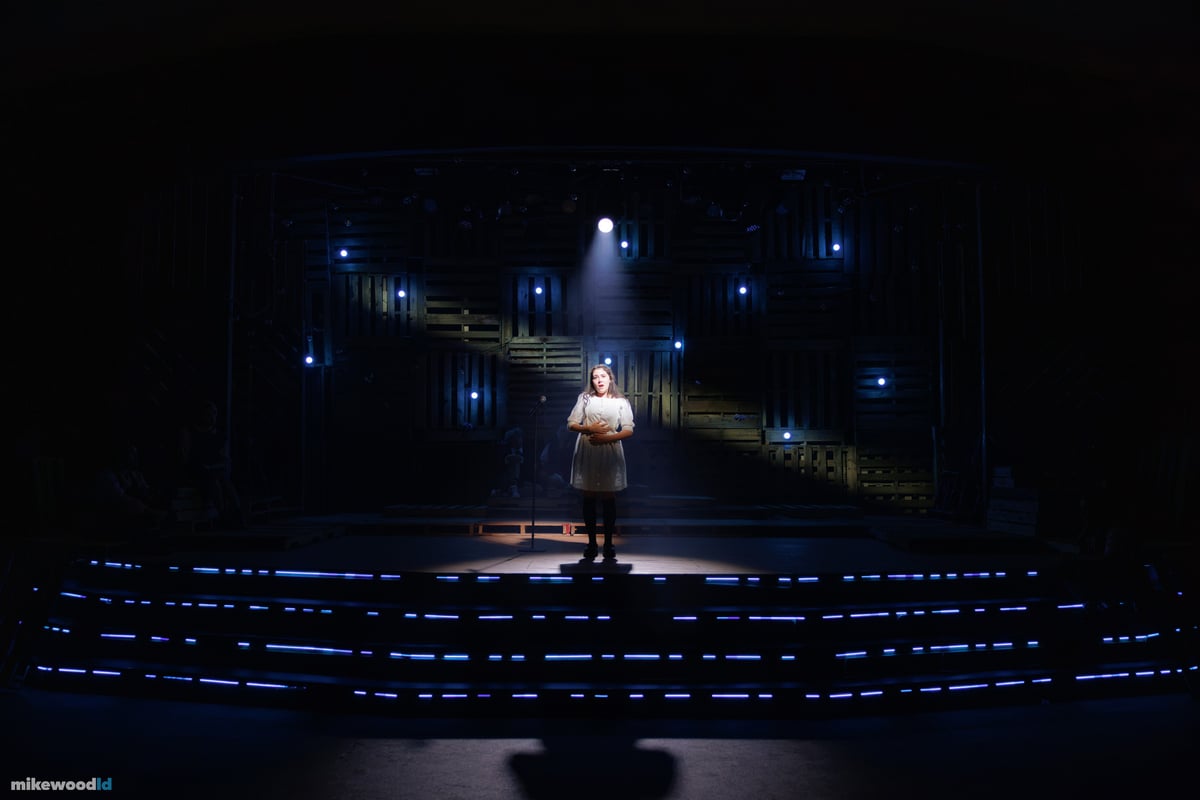 A young woman stands alone at a microphone in a narrow shaft of light on a near-black stage, faint blue practicals scattered across the pallet wall behind her.
