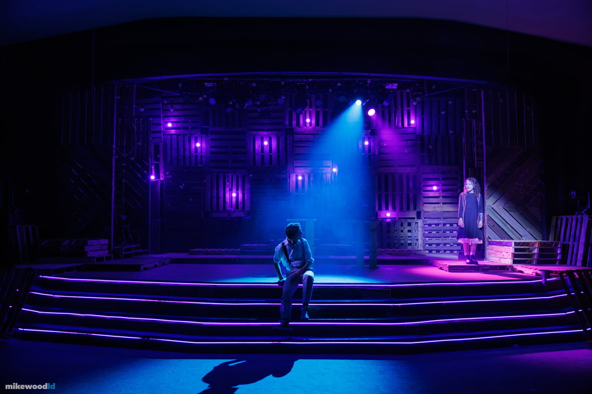 A young man sits alone on the glowing steps in a shaft of blue light and haze as a young woman watches from a platform at stage right, magenta practicals dotting the pallet wall.