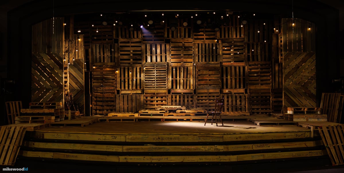 The empty pallet-wood set before the house opens, a single wooden chair centered on the plank stage under warm work lights and strings of bulbs.