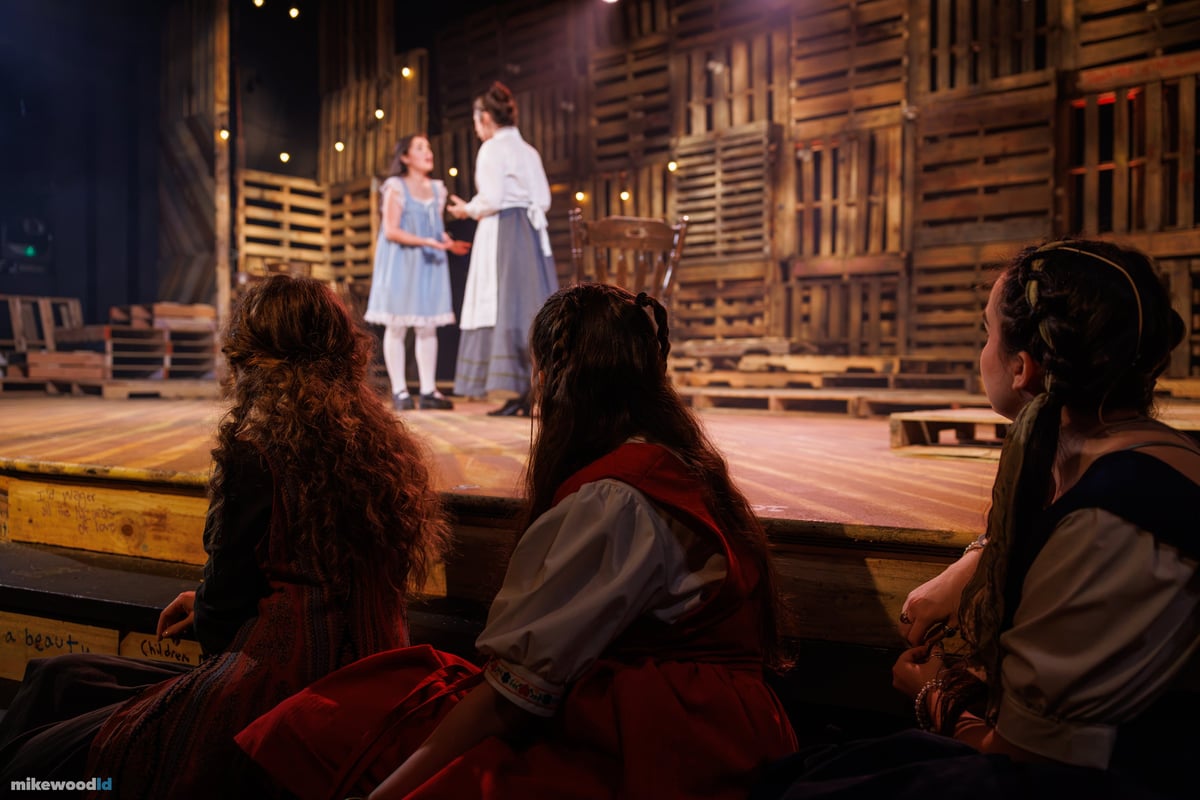Two actors in period costume speak on the warmly lit stage while three girls watch from the graffiti-marked front steps in the foreground.