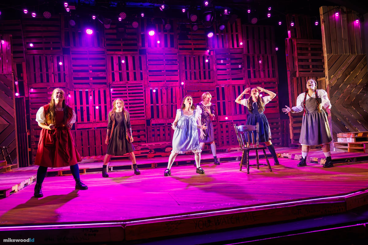 Six young women in period dresses sing spread across the stage in a saturated magenta wash, the pallet-wood wall glowing pink behind them.