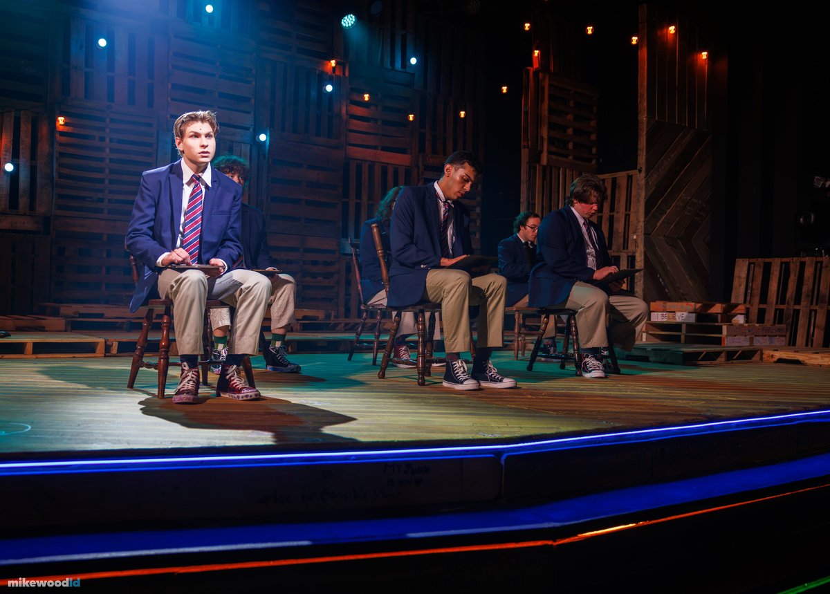 A schoolboy in a navy blazer and striped tie sits on a wooden chair with a book on his knee, classmates seated behind him in cool teal light with blue striping edging the steps.