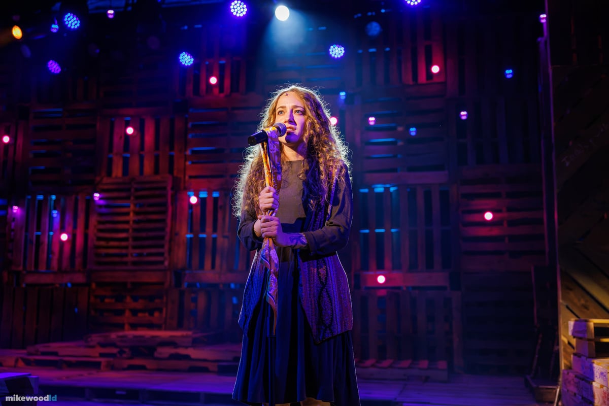 A young woman sings at a microphone stand under a blue and purple wash, colored pinlights scattered across the pallet-wood wall behind her.