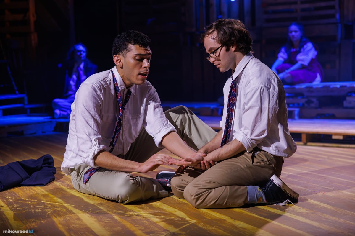 Two schoolboys in white shirts and ties sit close on the plank floor, one cradling the other's hand in soft blue light.
