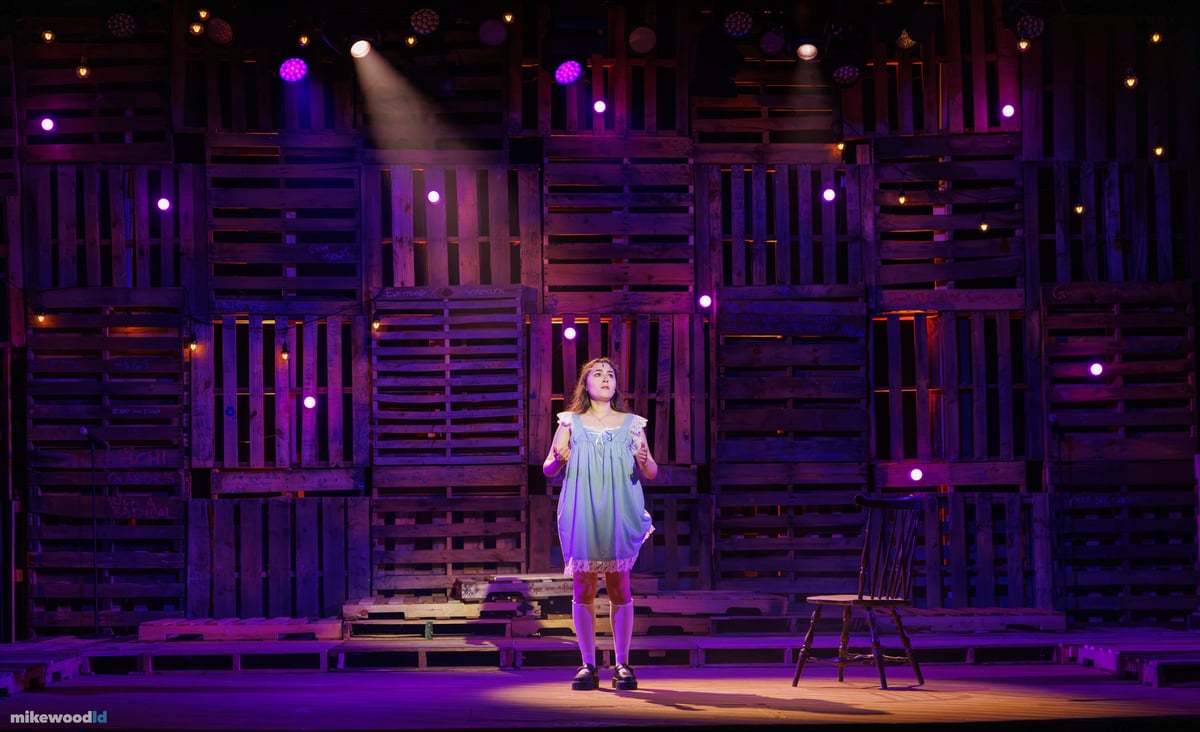 A young woman in a pale slip dress stands alone in a single top light within a deep violet wash, a wooden chair beside her and pinlights dotting the pallet wall.
