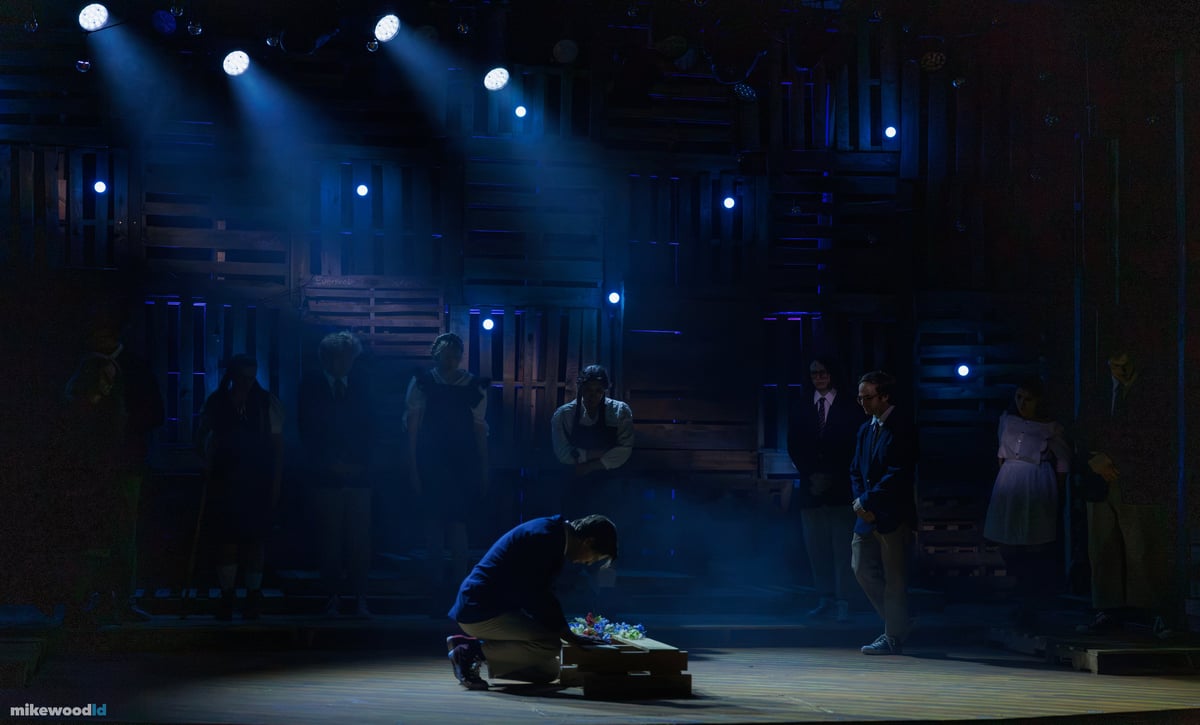 A lone figure kneels to lay flowers on a low crate while the company stands in a dim ring of silhouettes through smoky blue light.