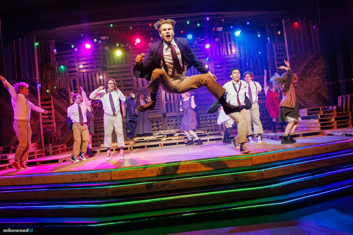 A schoolboy in a blazer and tie leaps high with a microphone as the company dances across the steps under colorful pinlights, neon striping edging the treads.