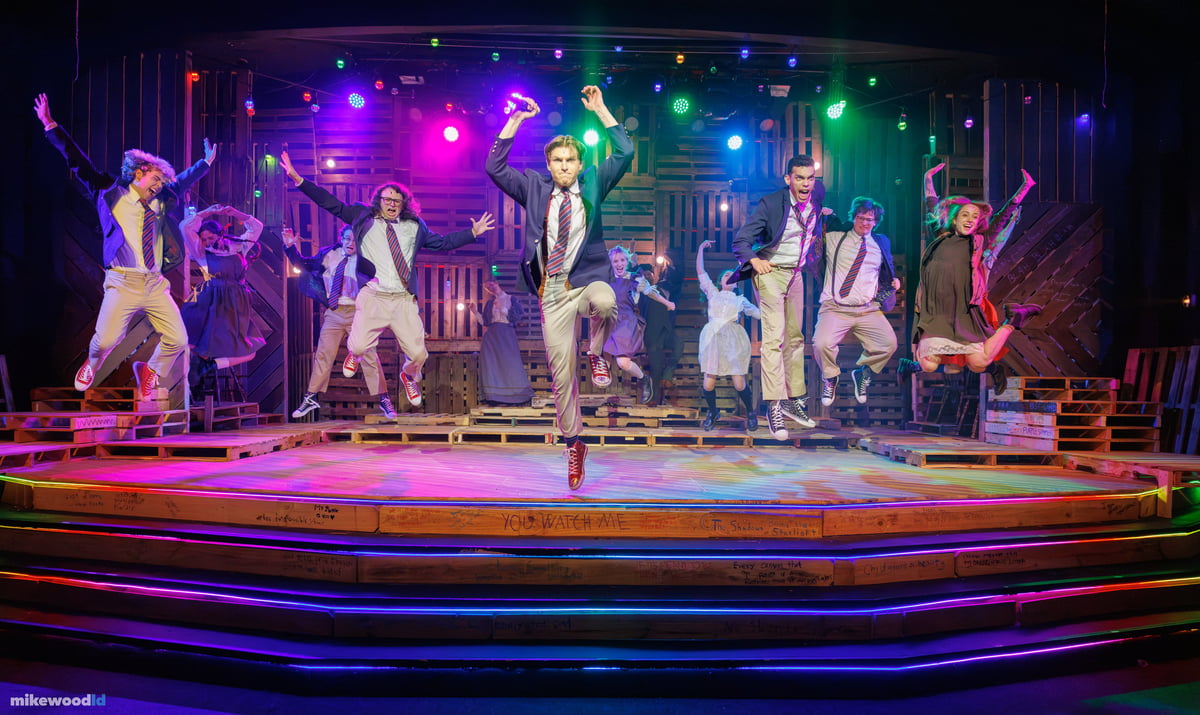 The whole company caught together mid-leap over the pallet stage, arms flung overhead, the step edges glowing in rainbow neon.
