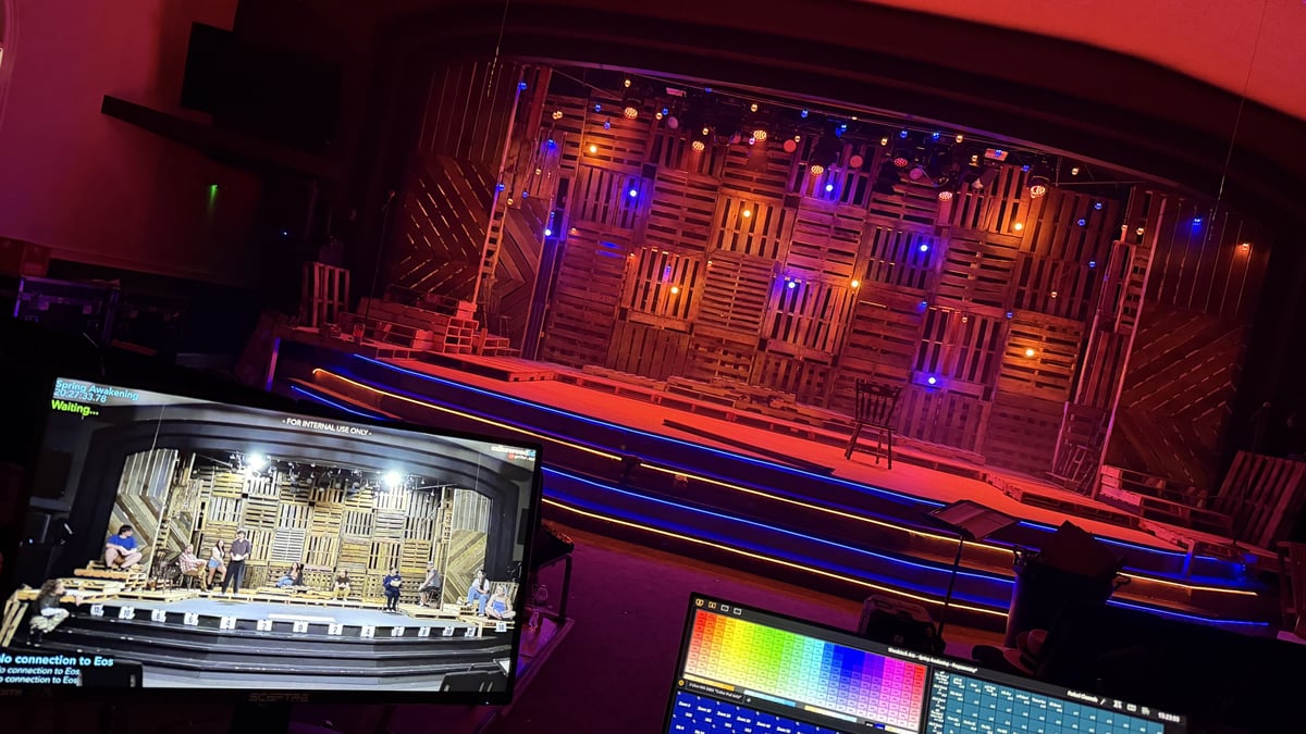 View from the tech table: the lit pallet-wall set beside a stage-feed monitor and the Eos programming screen