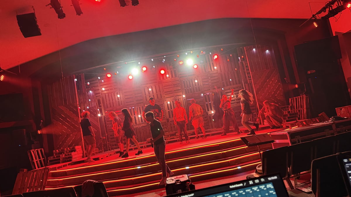 The cast rehearsing on the pallet-wall set under a red wash, seen from the booth