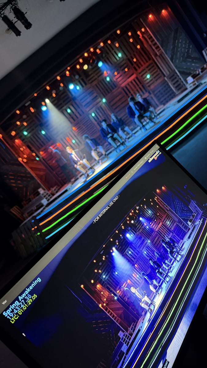 Booth view during tech, a stage-feed monitor of the schoolboys seated in blue light with the Vor capture screen below and neon step edges glowing