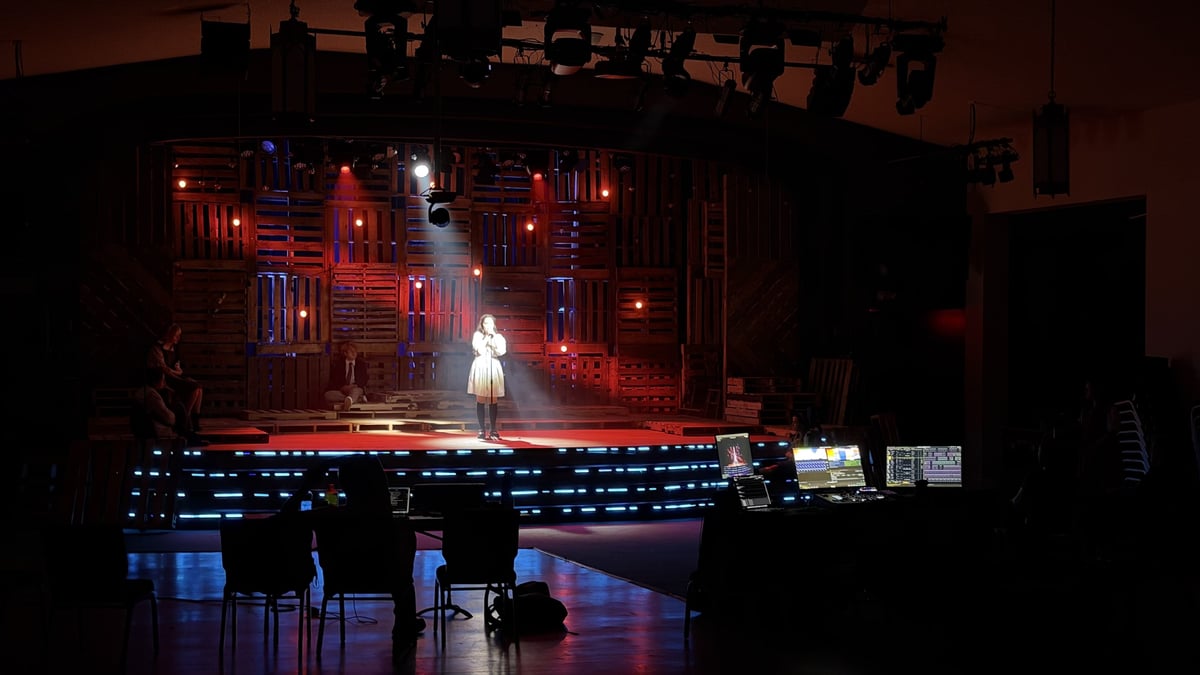 Wide view from the back of house, a lone actor spotlit on the set in a warm wash with the tech tables and empty seats in the foreground