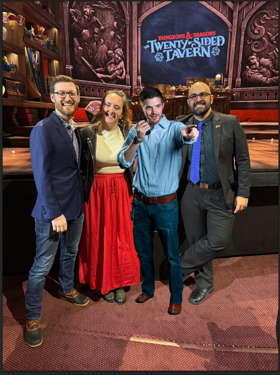 Four of the production team posing in front of the carved Twenty-Sided Tavern proscenium, each holding up a twenty-sided die.