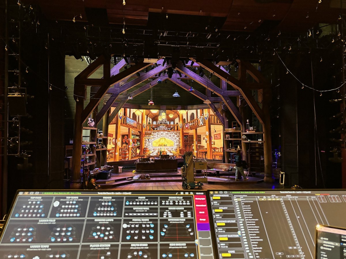 View from the front-of-house lighting console toward the warmly lit tavern set, Eos programming monitors glowing in the foreground.