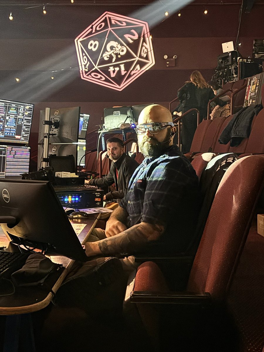 Two crew members at the tech table in the house with laptops and console monitors open, a glowing pink twenty-sided die suspended overhead.