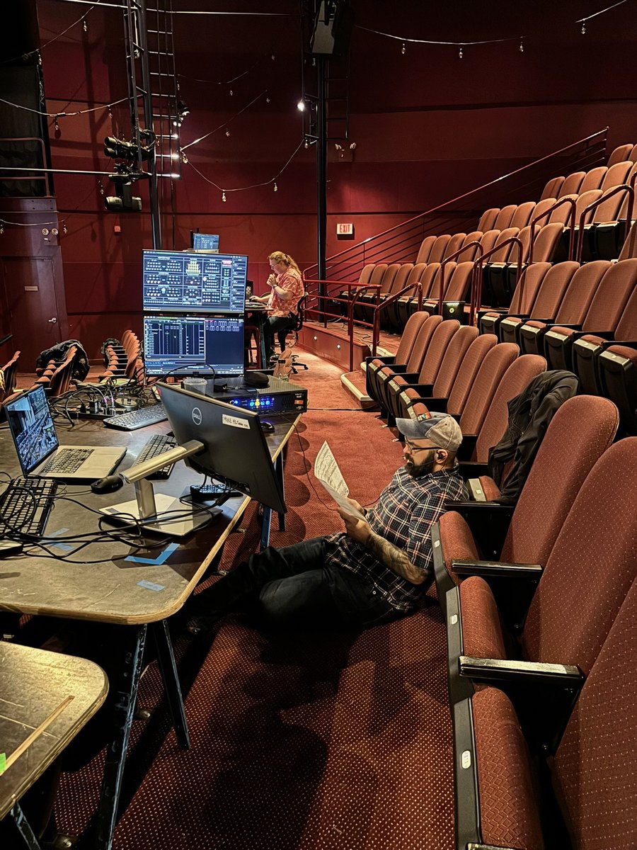 The tech table built across rows of house seats, lighting console and laptops on planks, one crew member reading paperwork on the floor between seats.