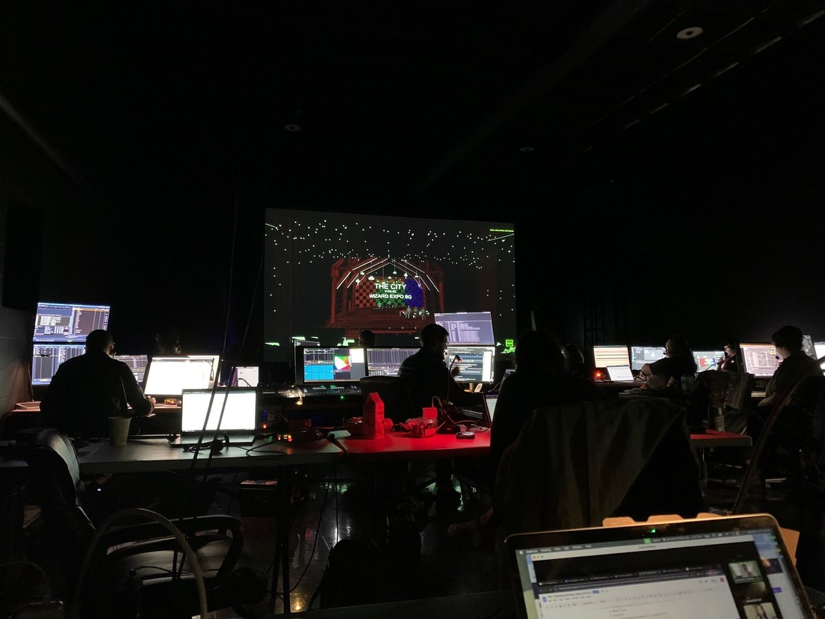 The darkened production table during tech, a row of glowing monitors with a city background plate on the projection screen behind.