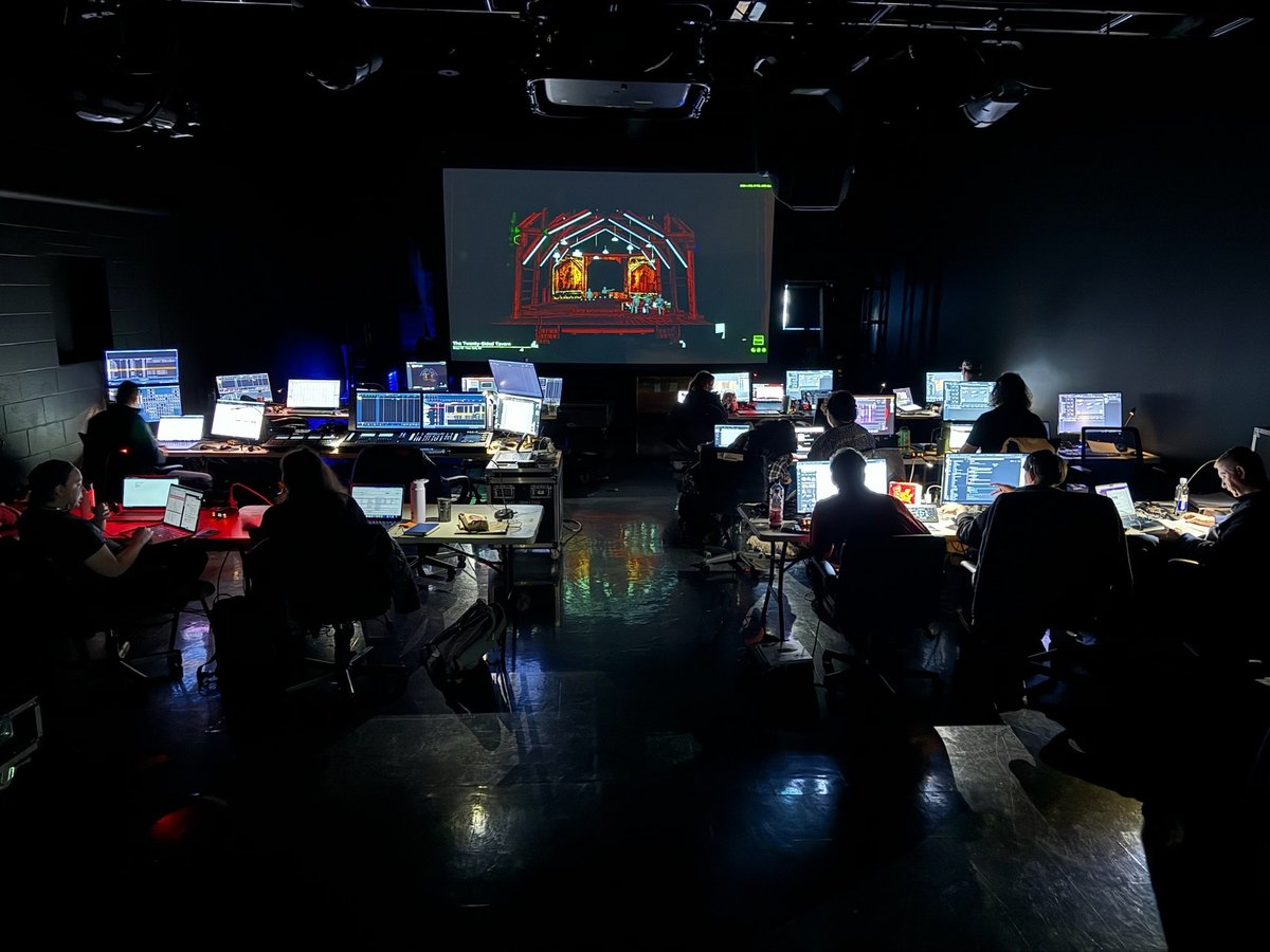 The full creative team at long tables of laptops in a dark production lab, a rendered model of the tavern set projected on the screen ahead.