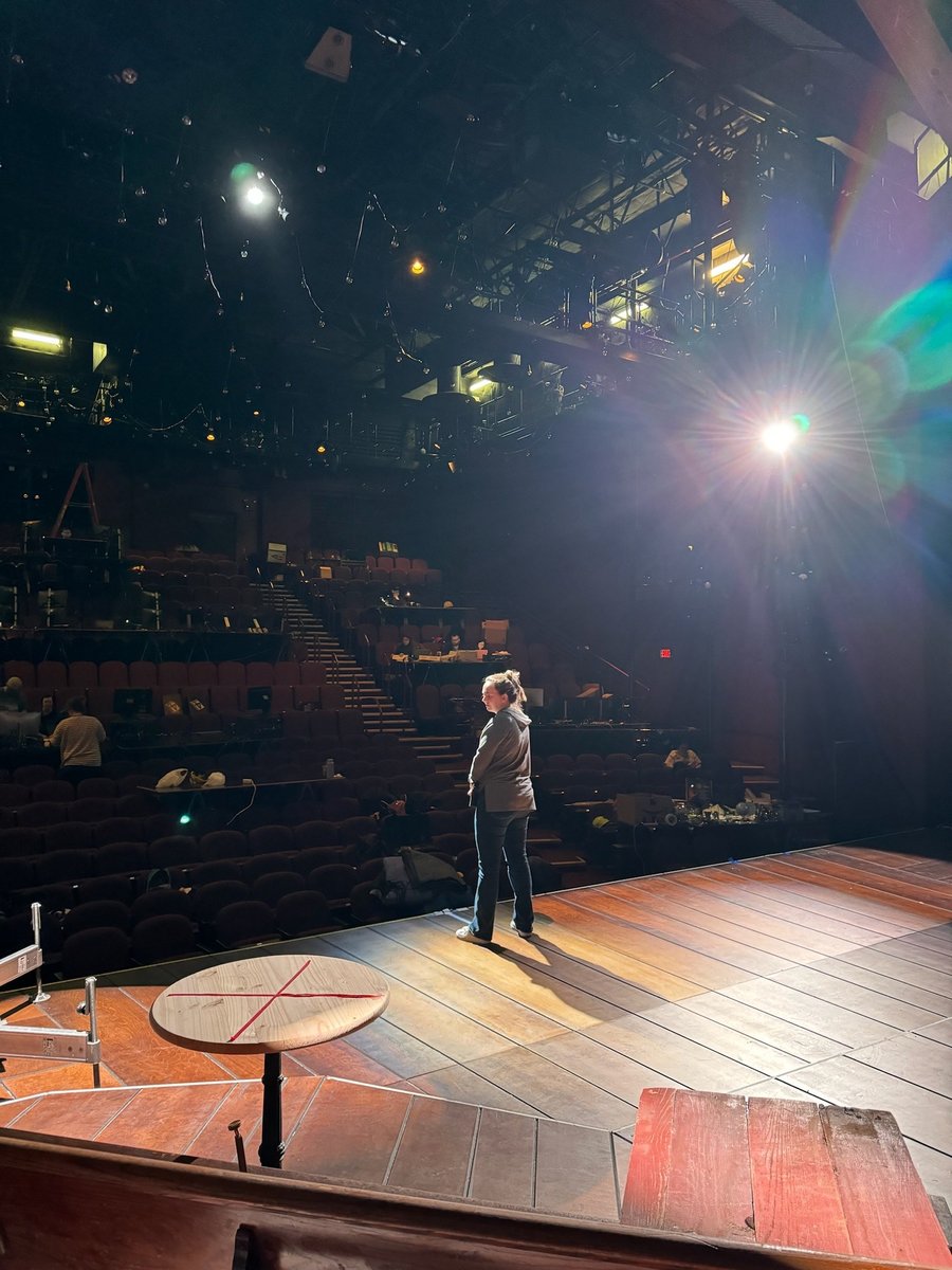 Looking out from the stage into the empty house during a focus session, a single fixture flaring bright overhead and a spike-marked table downstage.
