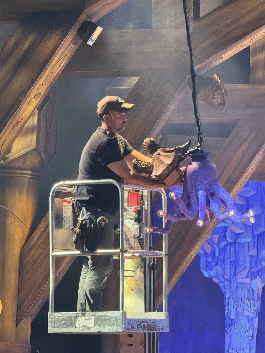 A technician on a personnel lift installing a large purple tentacled creature puppet into the tavern set amid atmospheric haze and blue light.