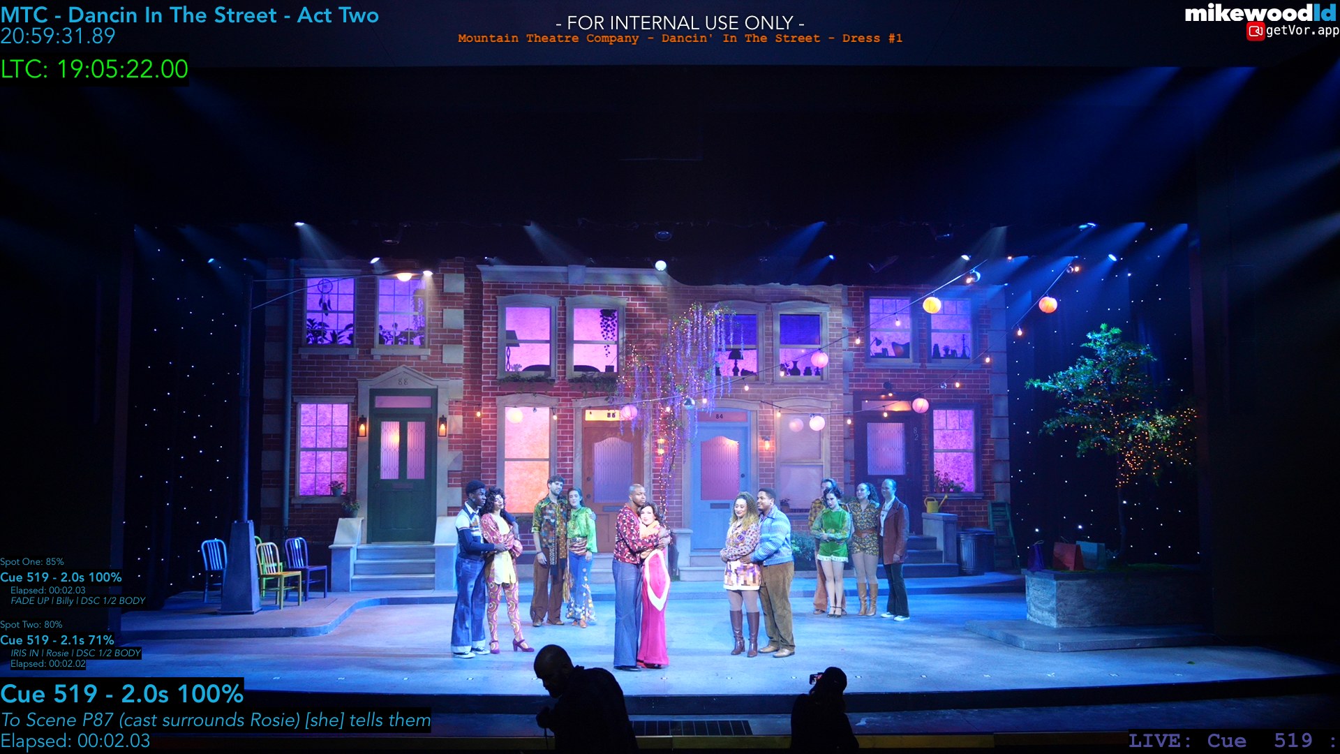 The same street at night mid-show: purple and magenta washes, a star-field sky, the cast gathered onstage