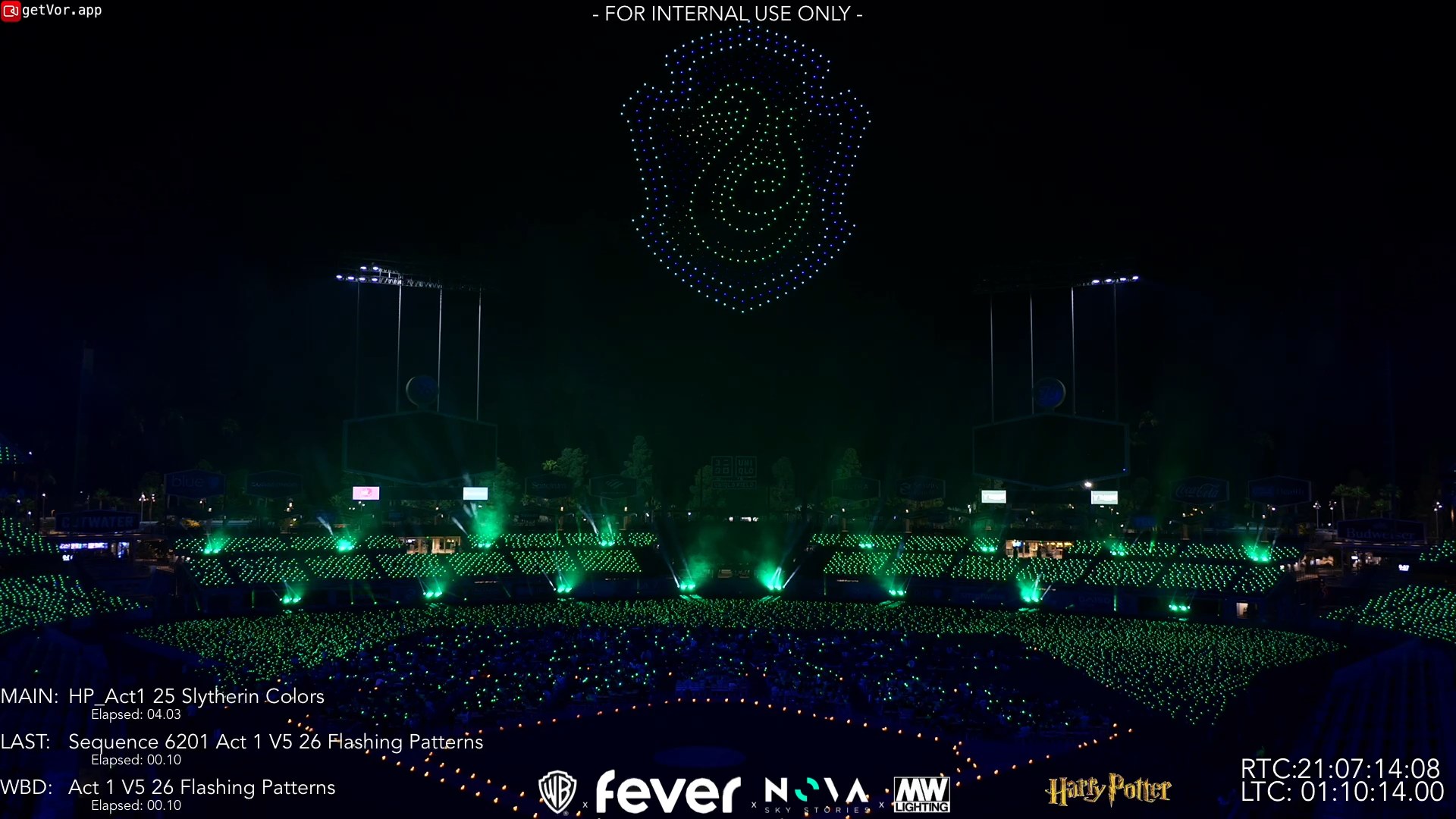 The same view at night: the Slytherin serpent drawn by drones above the field, green beams sweeping below