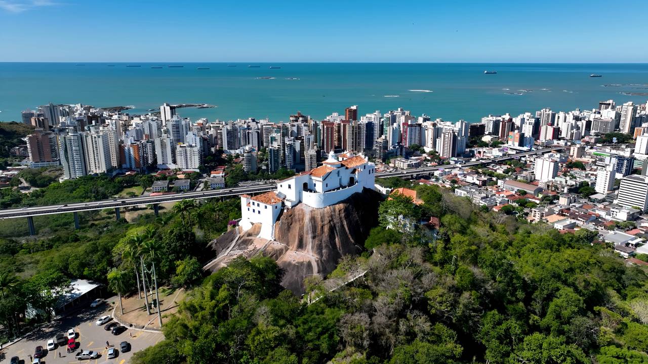Paisagem capixaba em destaque, com construção histórica branca sobre morro e mar ao fundo