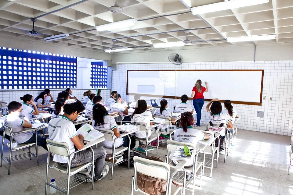 Professora em sala de aula da rede pública, escrevendo na lousa, com estudantes atentos