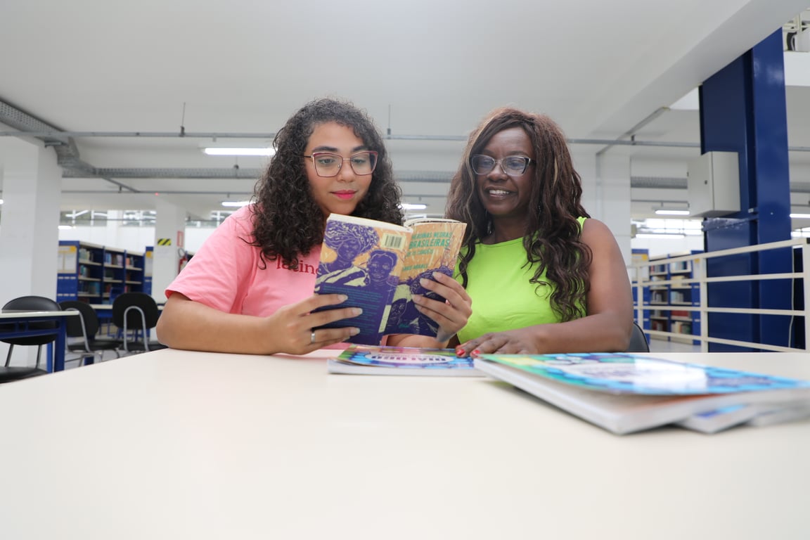 Ambiente acadêmico em biblioteca universitária; duas pessoas consultam um livro