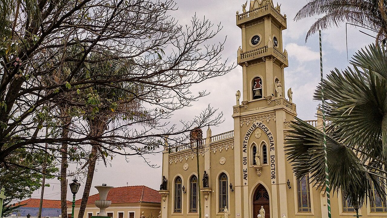 Vista aérea de cidade do interior paulista, com ruas arborizadas e casas baixas; ao fundo, igreja em destaque no cenário urbano