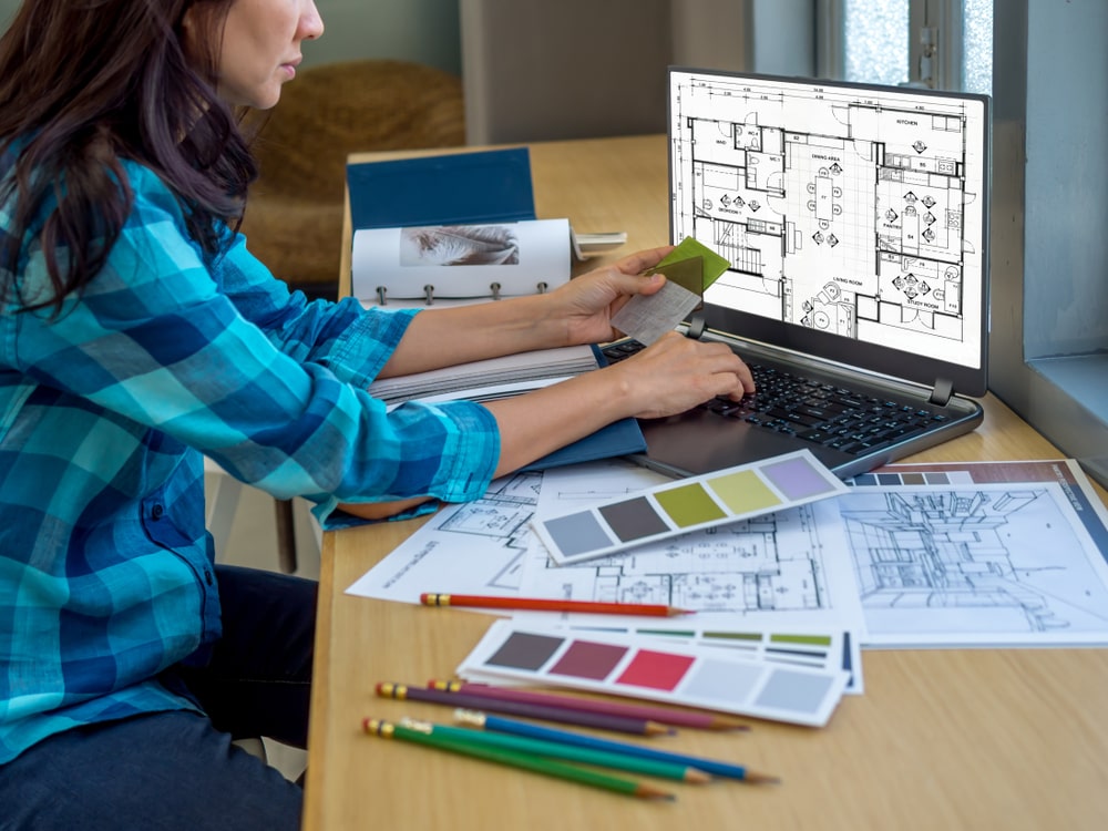 Foto de estúdio de arquitetura com uma pessoa trabalhando em um computador; mesa com desenhos e amostras de materiais.