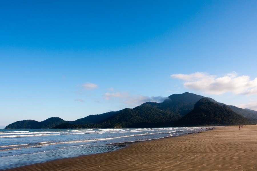 Praia de Peruíbe, SP, com montanhas ao fundo e céu ensolarado