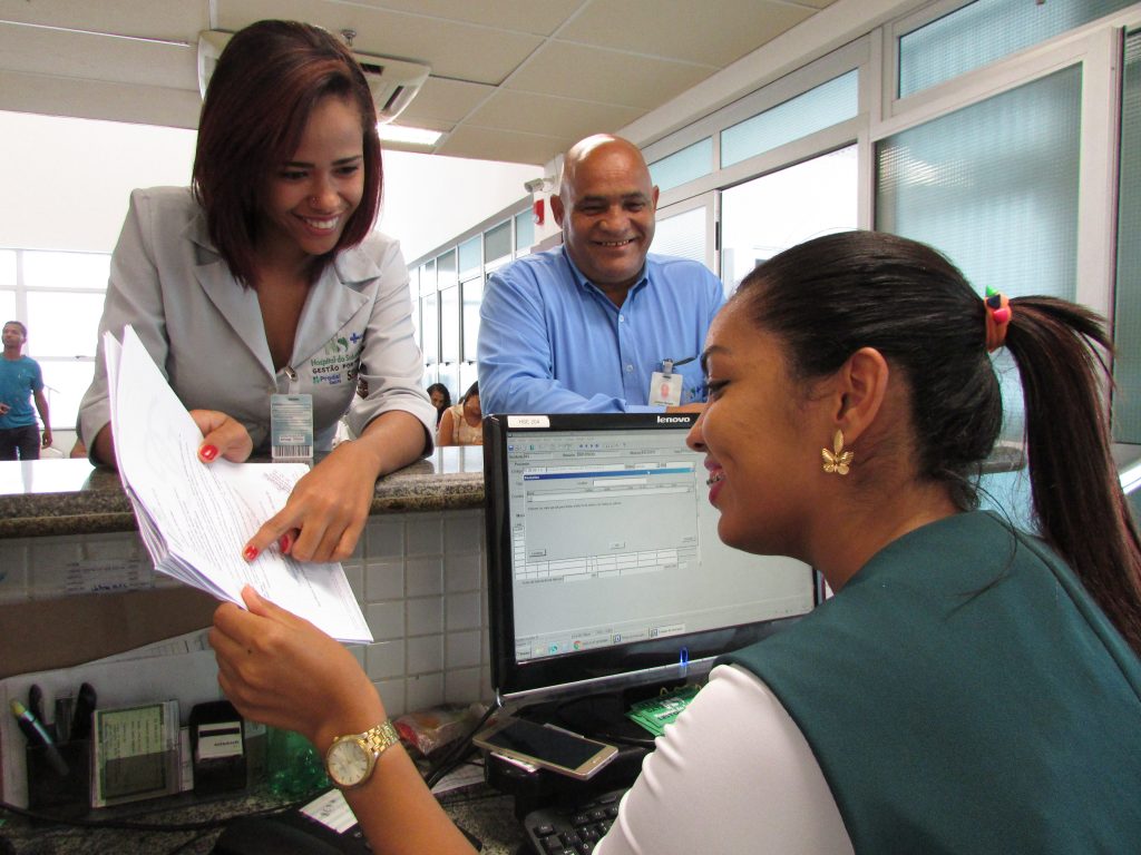 Recepção em ambiente hospitalar, com profissionais de saúde e administrativos atendendo ao público e operando computadores