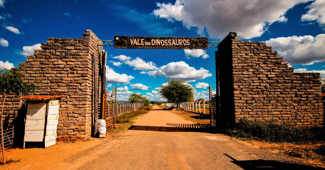 Entrada do Vale dos Dinossauros em Sousa PB sob céu azul, com visitantes ao fundo