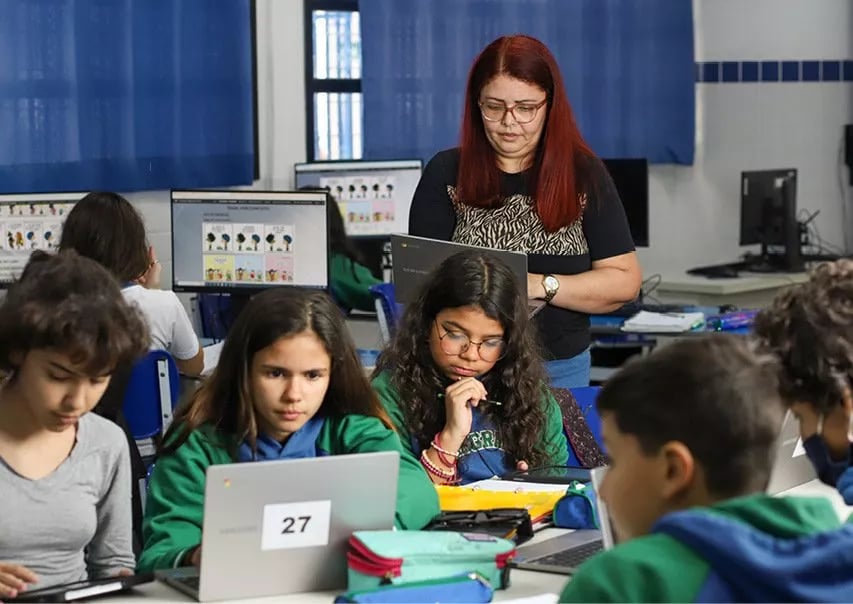 Sala de aula moderna com estudantes em computadores e professora acompanhando as atividades