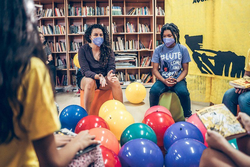 Sala de aula de EJA com adultos aprendendo, professora orientando alunos, ambiente iluminado e estantes de livros ao fundo