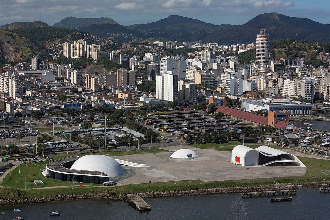 Vista aérea de uma cidade com prédios e áreas verdes. A paisagem mostra morros ao fundo e um céu claro.