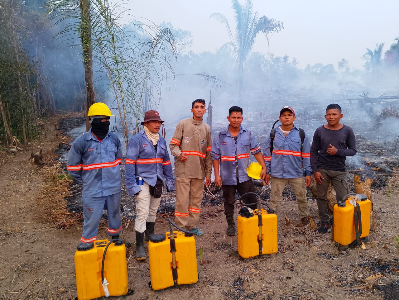 Brigadistas florestais equipados com EPI e bombas costais atuam próximos à fumaça em área de vegetação amazônica.