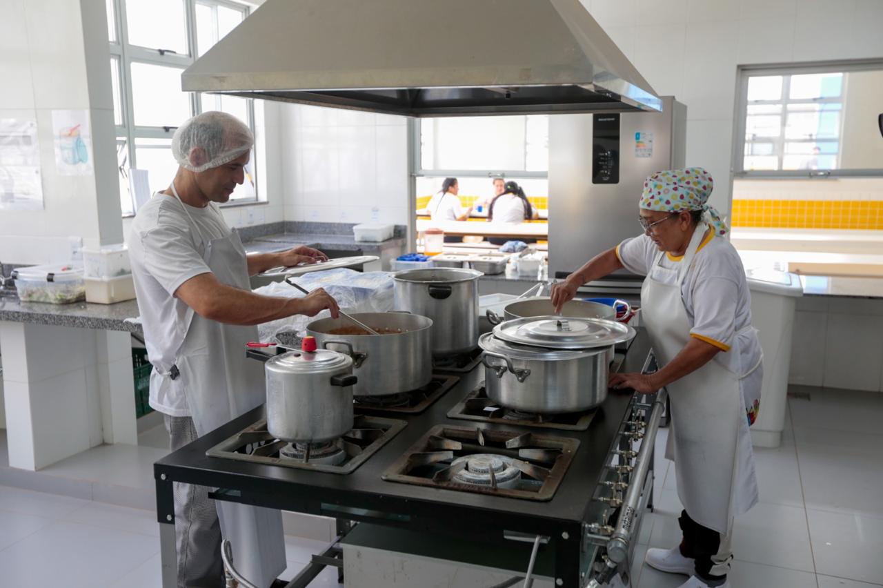Cozinha escolar com merendeiras preparando refeições em panelas grandes; ambiente limpo e organizado